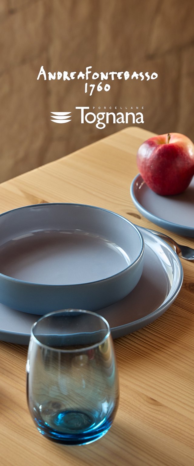 Set di stoviglie in porcellana blu su un tavolo di legno, composto da un piatto rotondo grande, un piatto più piccolo, un bicchiere con base blu e una mela rossa.