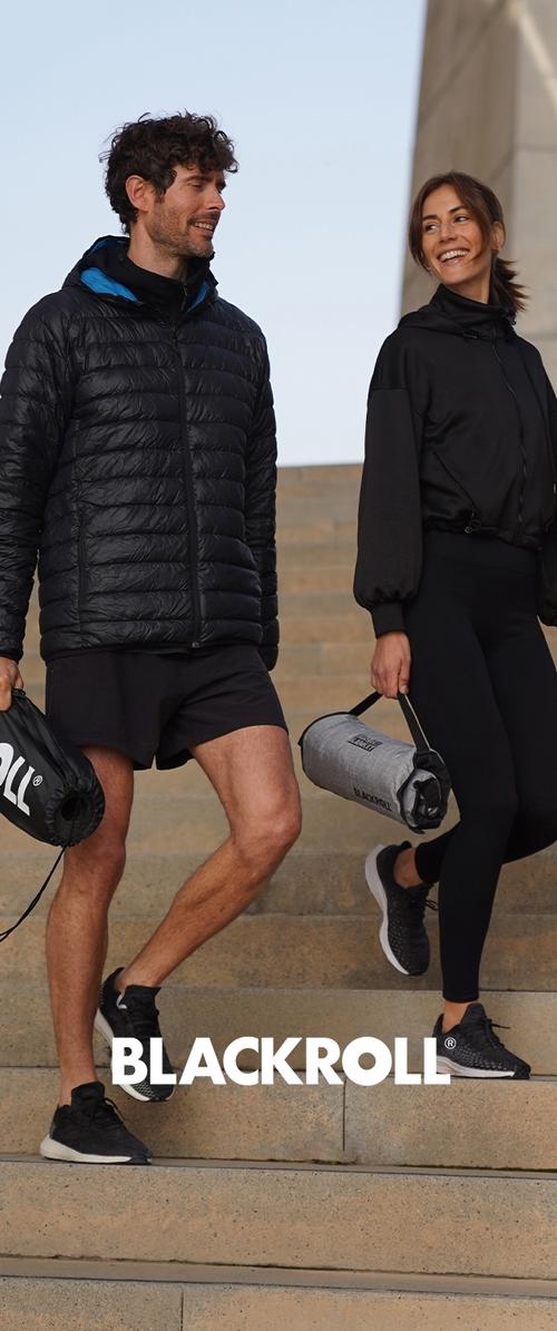 Zwei Personen steigen eine Steintreppe hinab, sie tragen schwarze Sportkleidung. Der Mann trägt eine schwarze, wattierte Jacke, die Frau ein eng anliegendes schwarzes Oberteil und Leggings.