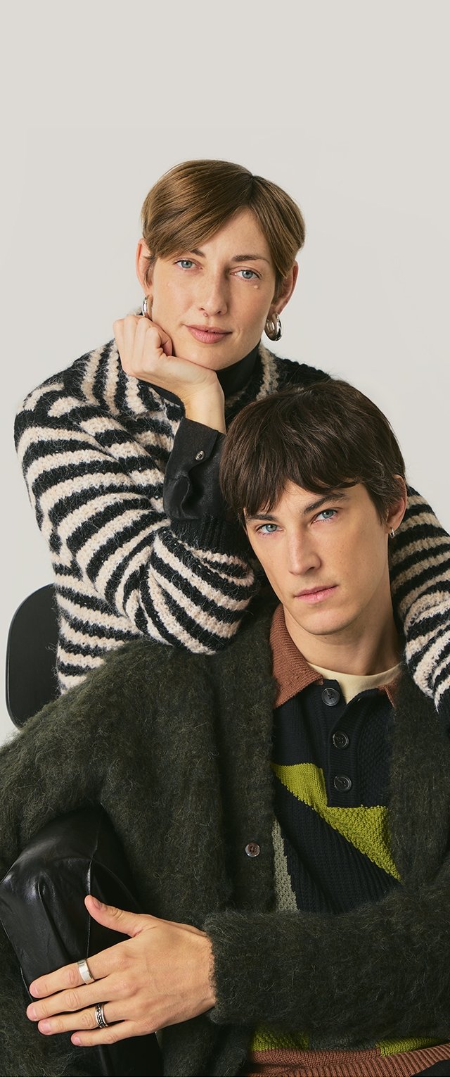 Deux modèles posent ensemble. La femme porte un pull rayé noir et blanc, tandis que l'homme a un cardigan vert sur une chemise en tricot multicolore.