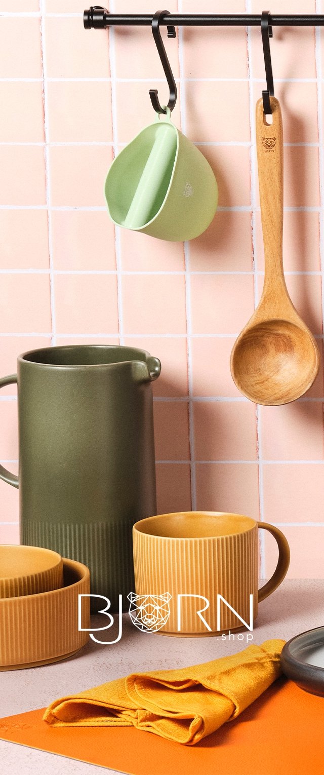 Keramikkrug in Olivgrün, gerippte orangefarbene Schalen und Tassen, Holzlöffel, grüner Messbecher und orangefarbene Serviette auf rosa gefliestem Untergrund.