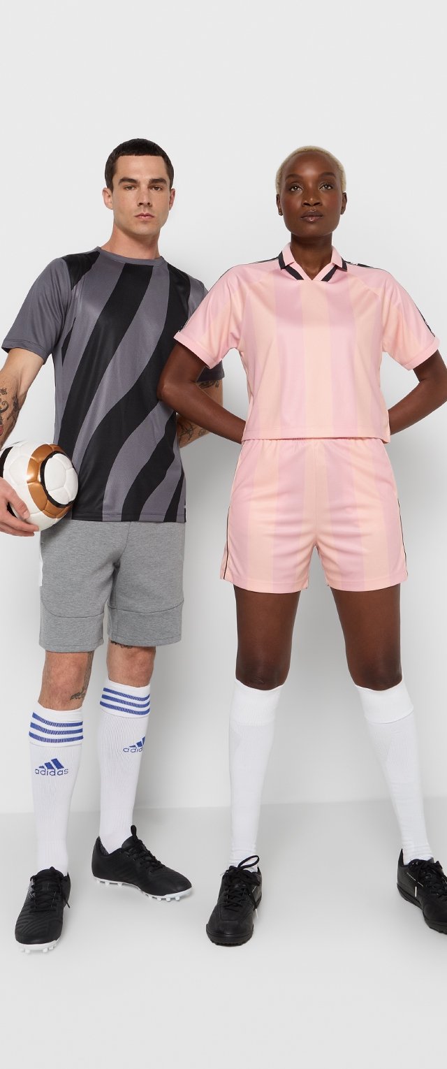 Camiseta deportiva a rayas grises y negras, pantalones cortos a rayas rosas, pantalones cortos grises, calcetines blancos con rayas azules y calzado negro. Se sostiene un balón de fútbol.