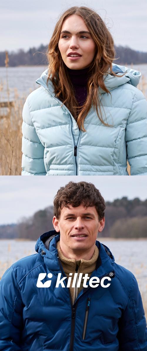 Femme en veste matelassée bleu clair et homme en veste matelassée bleu foncé se tiennent à l'extérieur près d'un lac avec des roseaux et des arbres en arrière-plan.