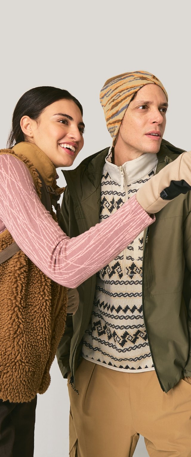 Smiling woman points ahead while standing next to a man wearing a patterned beanie and winter jacket, both dressed for cold weather.