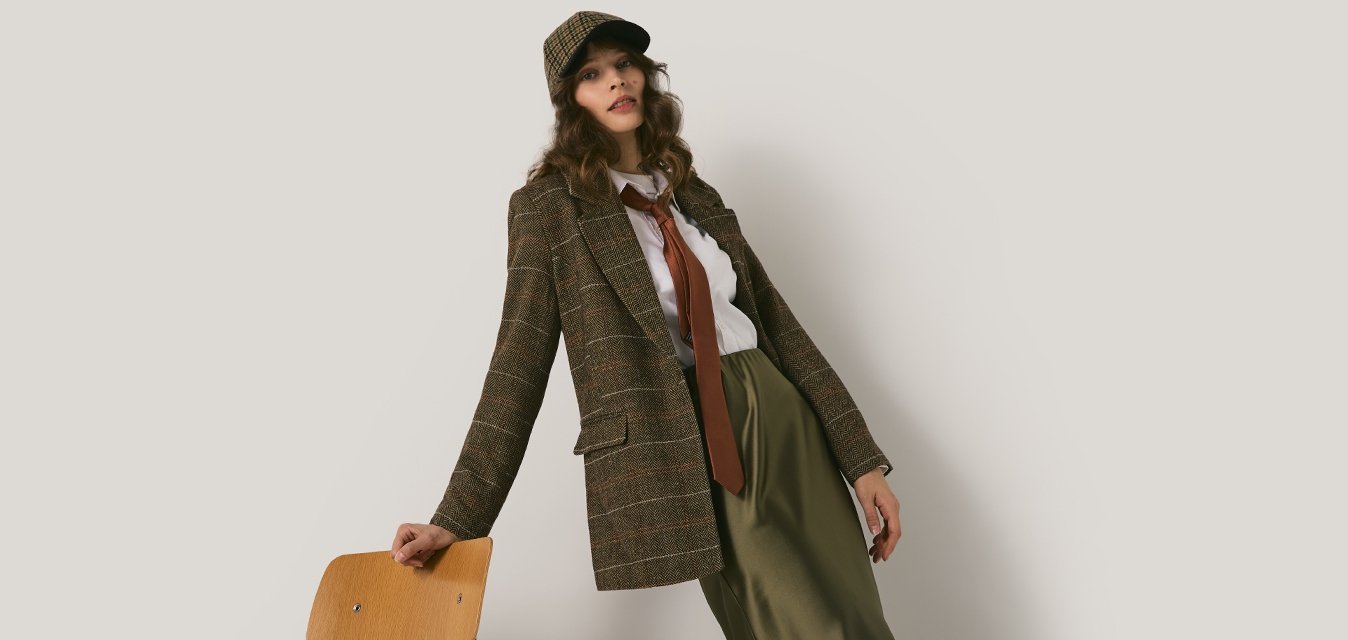 Žena v hnědém vzorovaném saku, odpovídající čepici, červené kravatě, bílé košili a zelených kalhotách se opírá o dřevěnou židli na světlém jednobarevném pozadí.