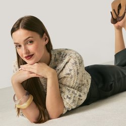 Žena leží na podlaze s bradou opřenou o ruce, má na sobě vzorovaný svetr, černé kalhoty, hnědé podpatky a náramky na jednoduchém pozadí.