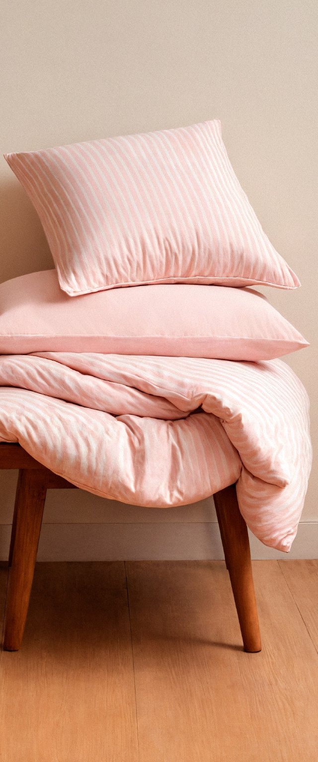 Juego de cama a rayas rosas exhibido en un banco de madera, incluye dos almohadas y un edredón con texturas suaves y colores pastel.