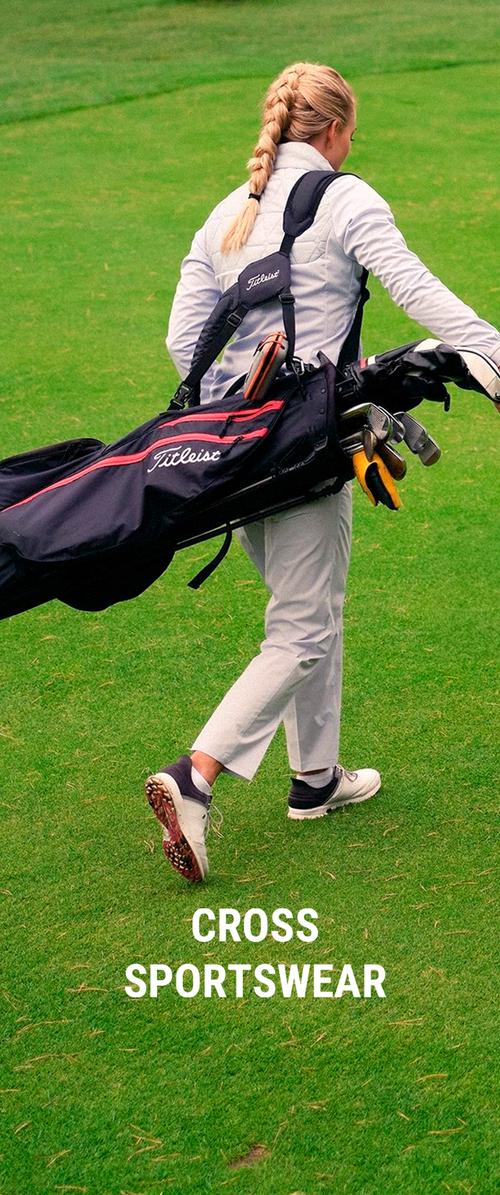 Person with braided hair walking on grass carrying a black and red Titleist golf bag with clubs, wearing white and gray sportswear.