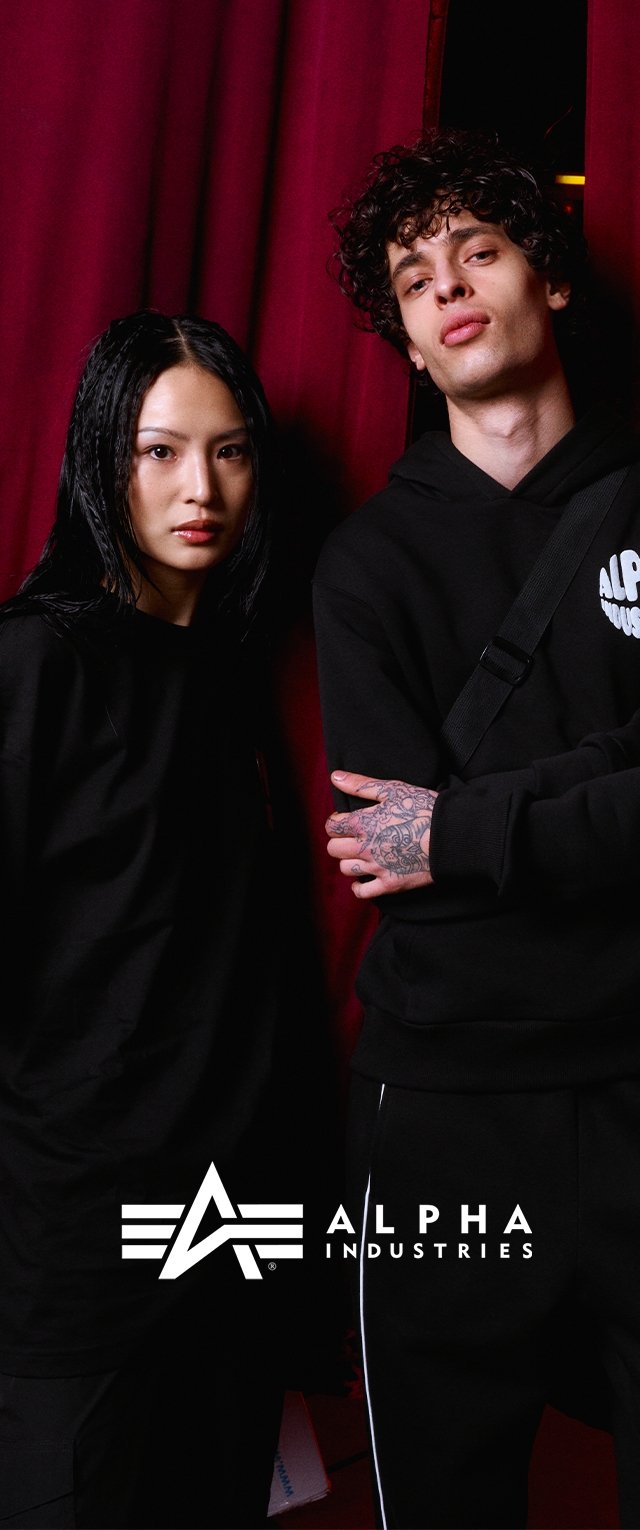 Two individuals wear black hoodies by Alpha Industries. One has long, wet hair; the other has curly hair and tattoos. The background is red.