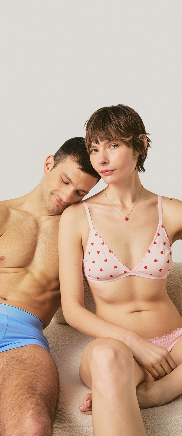 Man i blå underkläder vilar huvudet på axeln av en kvinna som bär en rosa BH och underkläder med hjärtmönster och sitter på en beige soffa.