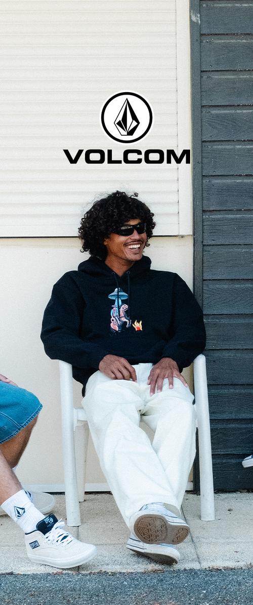 Young man with curly hair and sunglasses sits smiling on white chair wearing black hoodie and white pants outside a building with Volcom logo above.