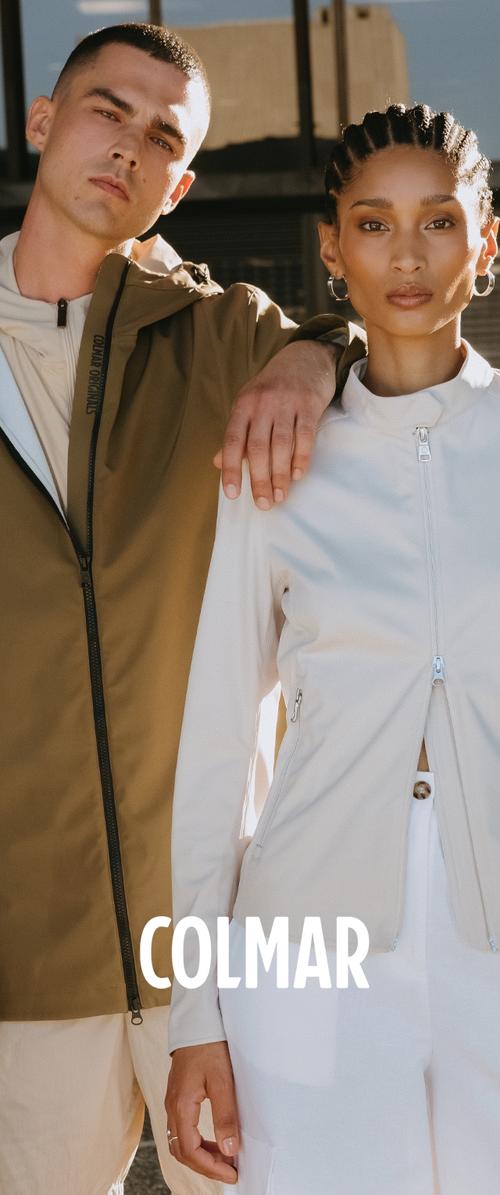 Man in brown jacket with zipper and woman in light-colored zip-up jacket stand close, man’s arm resting on woman’s shoulder outdoors.