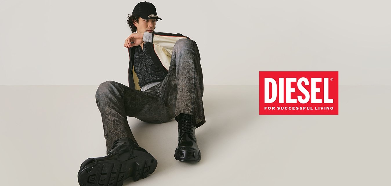 Osoba v černé čepici, černo-bílé bundě, šedých kalhotách a černých kozačkách sedí na podlaze proti jednobarevnému světlému pozadí.