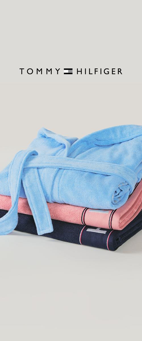 Hög med vikta handdukar i blått, rosa och marinblått med en blå morgonrock ovanpå under Tommy Hilfiger-logotypen på en ljus bakgrund.