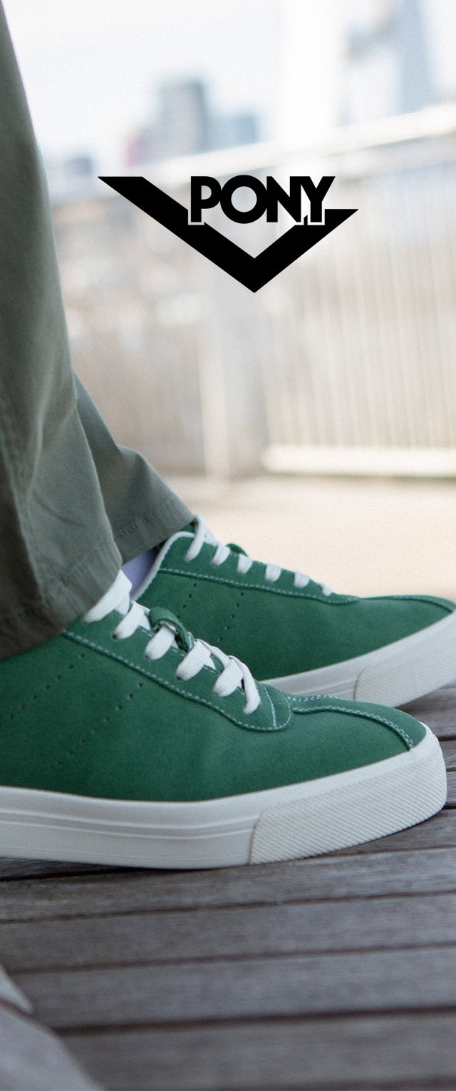 Baskets en daim vert avec des accents blancs, présentant un profil bas, un bout rond et des lacets blancs, posées sur une surface en bois. Logo visible.