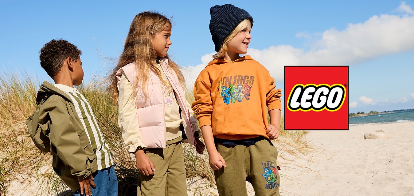 Tři děti stojí na písčité pláži poblíž písečných dun s trávou pod jasnou modrou oblohou, oblečené v neformálním podzimním oblečení.