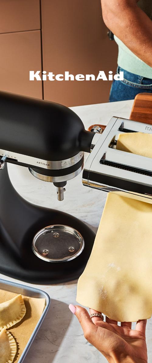 Person führt frischen Pastateig durch einen schwarzen KitchenAid Standmixer Pasta-Walzenaufsatz auf einer Marmorarbeitsplatte.