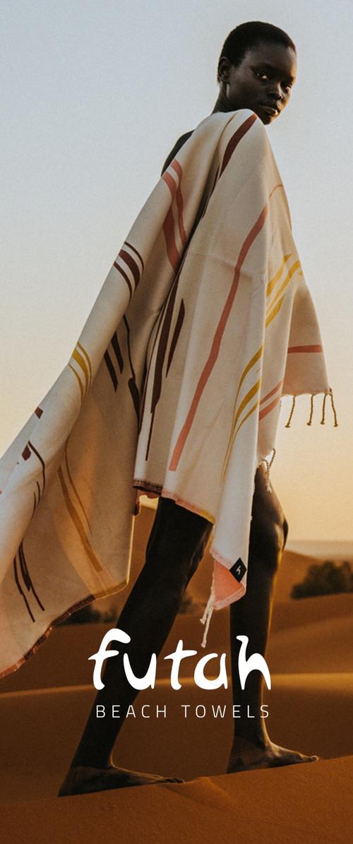 Persona de pie en dunas de arena al atardecer, envuelta en una gran toalla de playa futah estampada con rayas en tonos tierra.