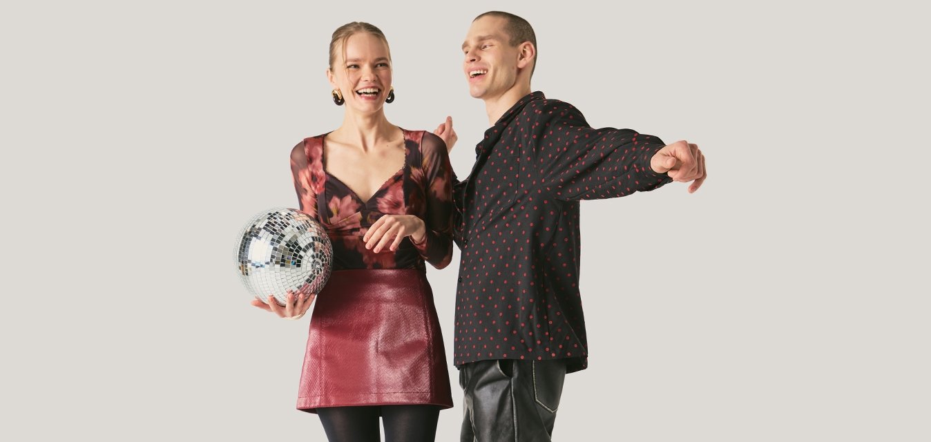 Žena držící disko kouli, oblečená v květovaném topu a červené sukni, směje se s mužem v černé vzorované košili a kožených kalhotách, kteří tančí.