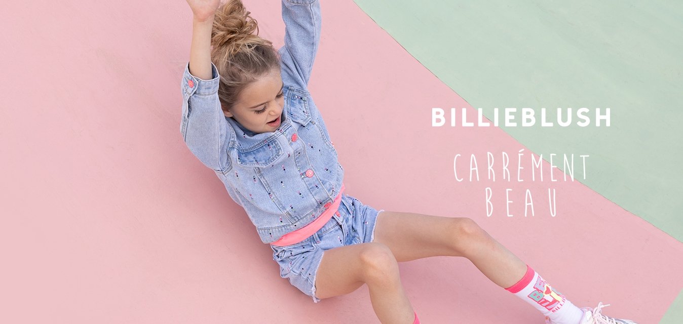 Mladá dívka v džínové bundě a krátkých kalhotách sjíždí zakřivený růžovo-zelený povrch hřiště s vytaženýma rukama.