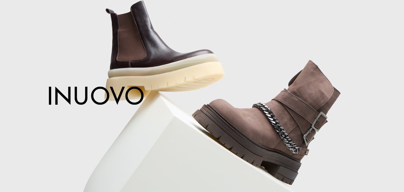 Dvoje kotníkových bot: jedny hnědé kožené s béžovou robustní podešví, druhé šedé semišové s řetízkovým detailem a černou gumovou podešví, vystavené na bílém podstavci.