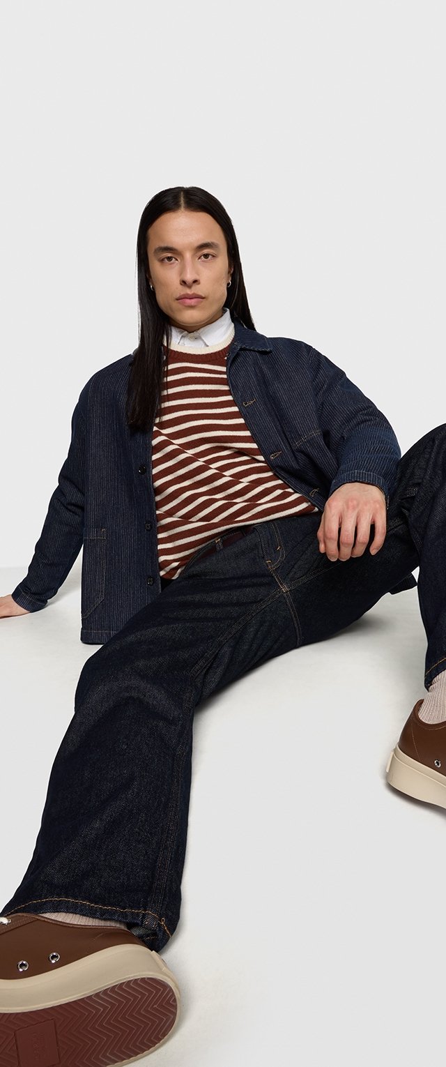 Jeansjacke, gestreifter Pullover in Braun und Creme, dunkle Jeans und braune Sneakers. Lange Haare, sitzende Pose mit ausgestreckten Beinen.