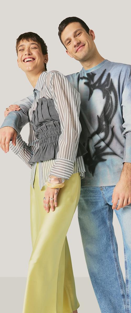Dos adultos sonrientes, uno con una camisa a rayas y falda amarilla, el otro con una sudadera con estampado y jeans, de pie muy juntos.