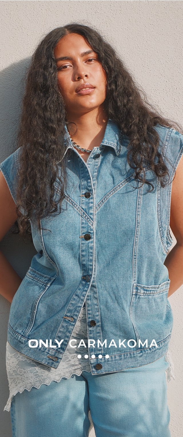 Persona con cabello largo y rizado que lleva una chaqueta vaquera sin mangas sobre un top de encaje y jeans azul claro, de pie contra una pared beige.