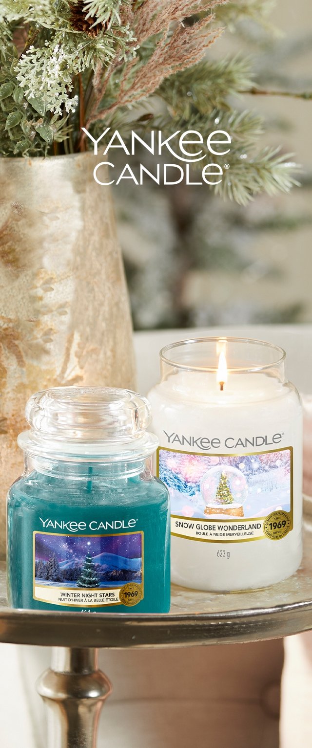Dos tarros de Yankee Candle: uno color azul verdoso etiquetado como "Estrellas de Noche Invernal" y otro blanco etiquetado como "Maravillas de Globo de Nieve" con una mecha encendida. Detalles y etiquetas doradas.