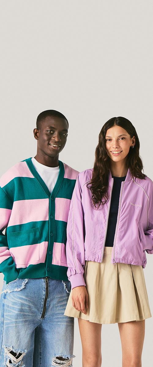 Jonge man in een gestreepte cardigan en gescheurde spijkerbroek staat naast een glimlachende vrouw in lila jas en beige rok tegen een effen achtergrond.