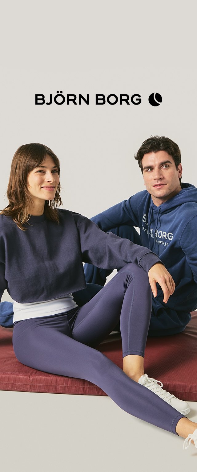 Donna sorridente in felpa blu navy corta e leggings seduta accanto a un uomo con felpa con cappuccio blu Björn Borg su un tappetino rosso con il logo del marchio sopra.