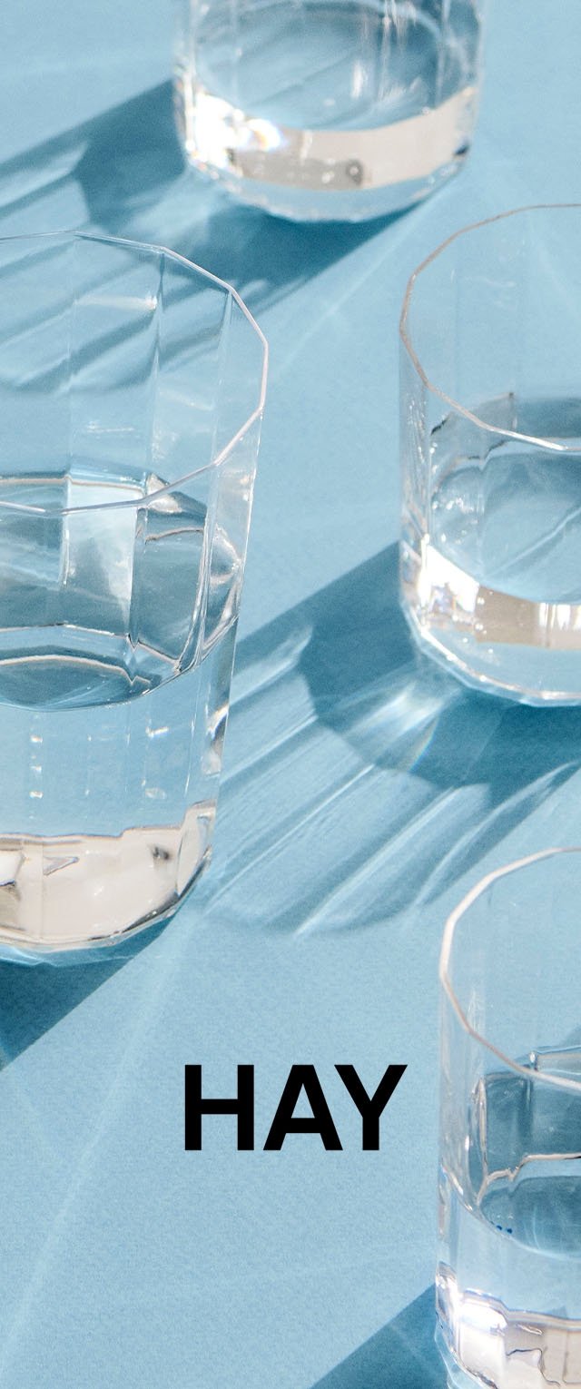 Vier facettierte Gläser aus klarem Glas werfen Schatten auf eine blaue Oberfläche, auf der der schwarze Schriftzug „HAY“ zu sehen ist.