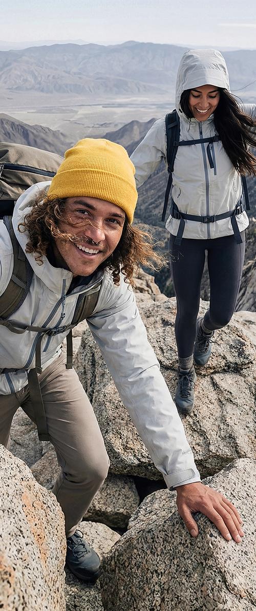 Due escursionisti con giacche leggere arrampicano su un terreno roccioso in un paesaggio montano, uno sorride alla telecamera e l'altro è concentrato sull'arrampicata.