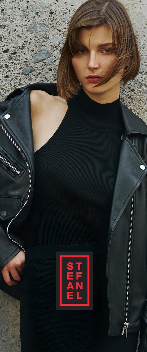 Chaqueta de cuero negra sobre un vestido de punto negro acanalado, con un escote alto y una etiqueta cosida prominente con letras rojas y negras.
