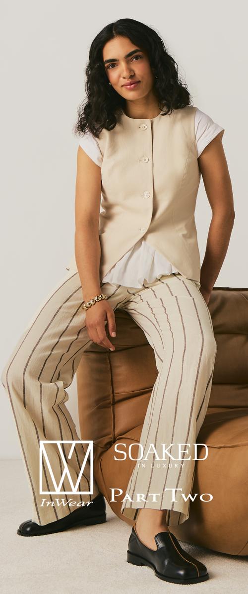 Kvinde med krøllet mørkt hår iført en beige vest med knapper, hvid skjorte, brede stribede bukser, sorte sko og et armbånd, der sidder på en brun sofa.