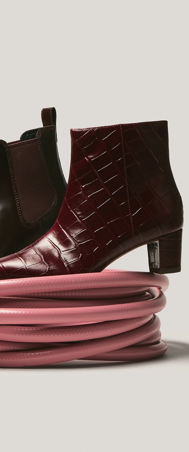 Burgundy ankle boots with a glossy crocodile texture and a block heel, paired with a pink coiled hose in the foreground.