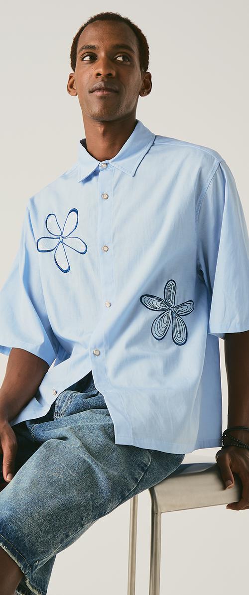 Man draagt een lichtblauw shirt met korte mouwen met donkerblauwe bloemmotieven en denim shorts, zittend op een witte kruk tegen een effen achtergrond.