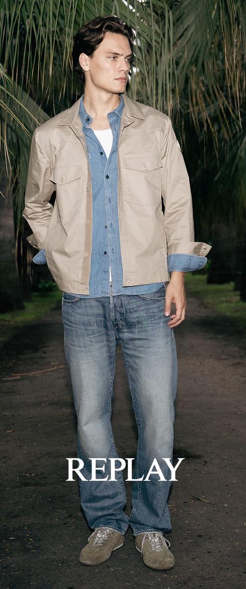 Joven con chaqueta beige sobre camisa de mezclilla azul y jeans desgastados, de pie en un camino de tierra con palmeras de fondo, texto "REPLAY" debajo.
