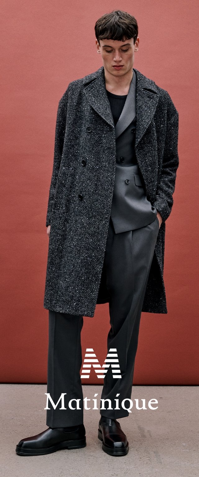 Jeune homme en costume croisé gris, chemise noire, pardessus gris à motifs, chaussures noires, debout devant un fond orange brûlé.