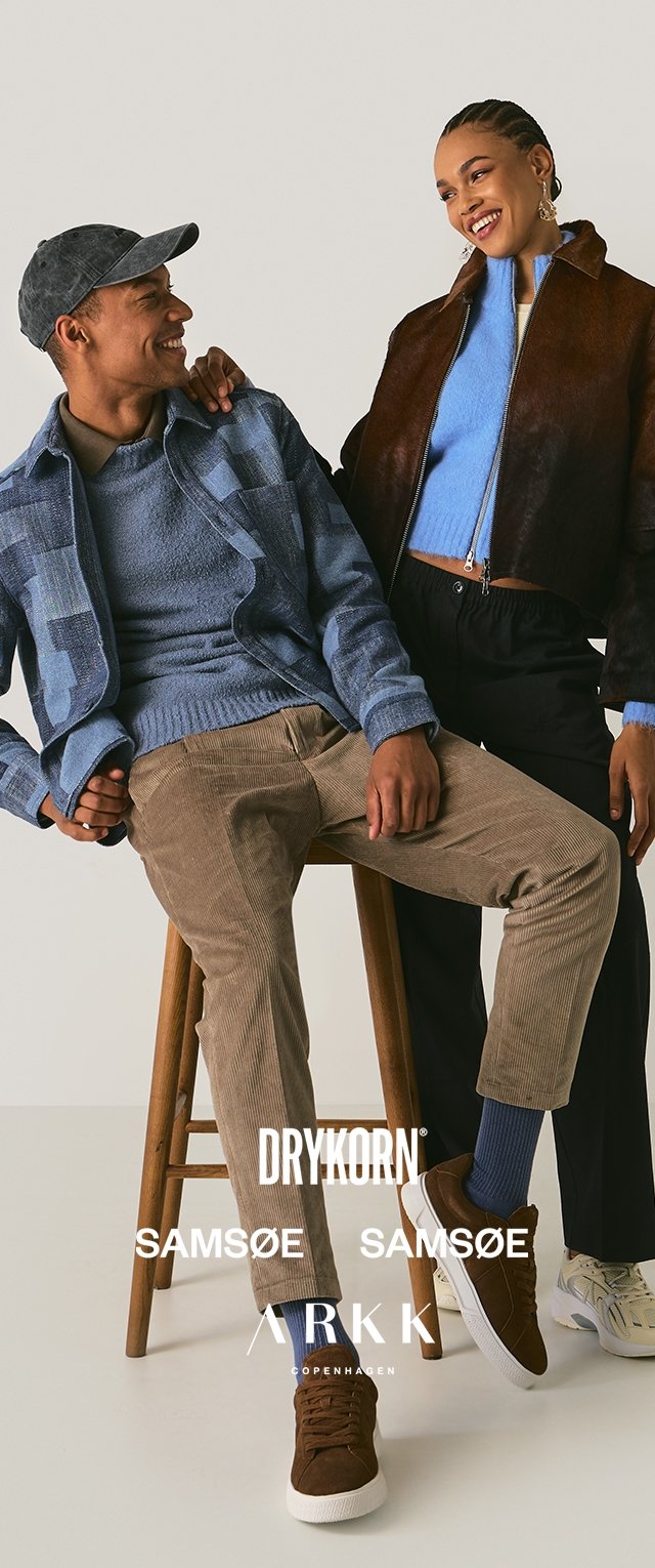 Twee mensen poseren op een houten kruk. De ene draagt een blauw geruit jasje, een blauwe trui en tan kleurige corduroy broeken. De ander draagt een bruin leren jasje, een lichtblauwe top en zwarte broeken. Beide dragen casual schoenen.