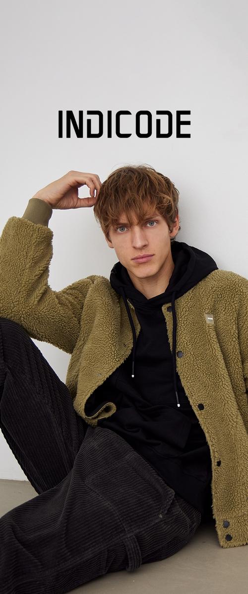 Veste en fausse fourrure vert olive portée sur un sweat à capuche noir, associée à un pantalon en velours côtelé foncé. Le modèle pose avec une main dans les cheveux, devant un fond neutre.