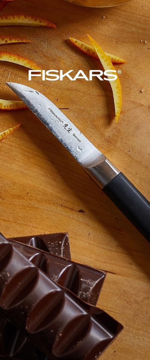 Cuchillo con mango negro y gotas de agua cerca de rodajas de naranja pelada y chocolate oscuro roto sobre una superficie de madera con el logo de Fiskars.