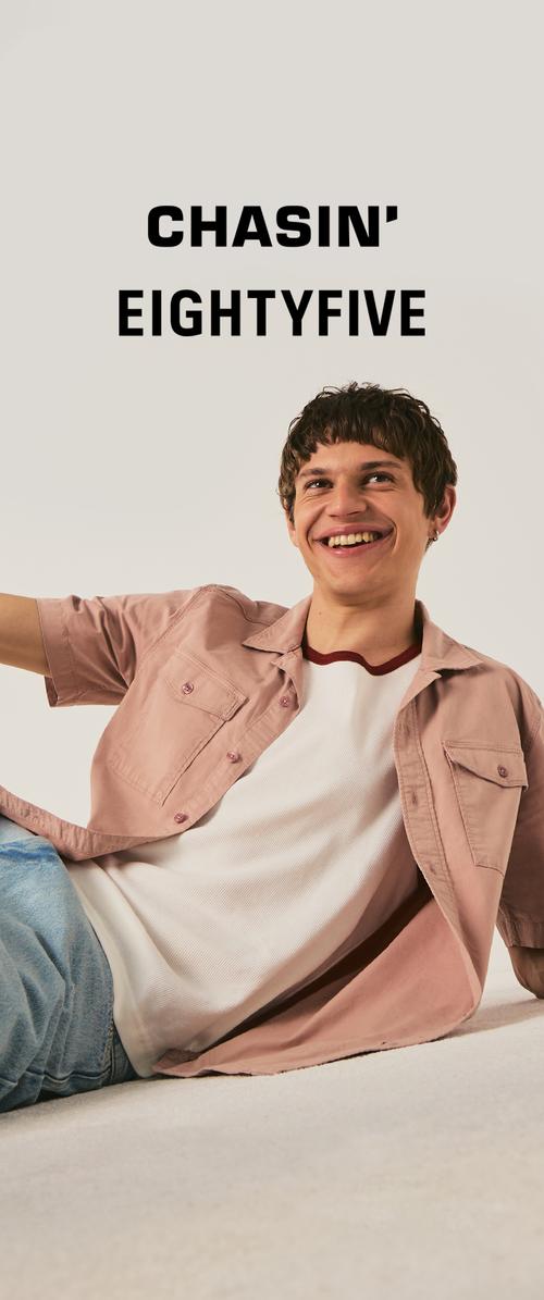 Jeune homme souriant aux cheveux courts et bouclés, portant une chemise rose clair sur un t-shirt blanc et un jean bleu, assis de manière décontractée devant un fond uni.