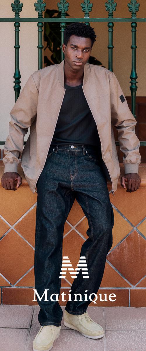 Jeune homme portant une veste beige, une chemise noire, un jean foncé et des chaussures claires, appuyé contre un mur carrelé surmonté d'une rambarde en fer vert.
