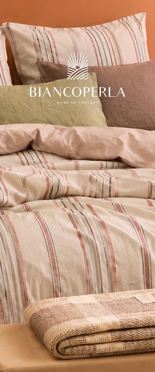 Ropa de cama a rayas en tonos crema y rosa empolvado, con almohadas superpuestas y una manta beige doblada, colocada sobre una cama; encima aparece la marca Biancoperla.