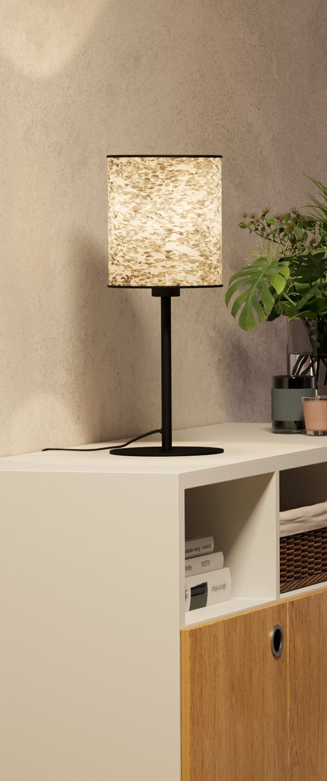 Lámpara de mesa encendida con pantalla beige texturizada sobre estante blanco junto a planta de hojas verdes en jarrón de vidrio y libros apilados en compartimentos.