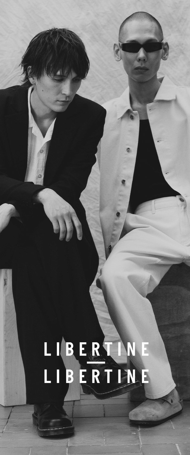 Zwei Models sitzen nebeneinander. Das eine trägt einen schwarzen Anzug und Stiefel; das andere eine helle Jacke und weiße Hose mit Slippern.