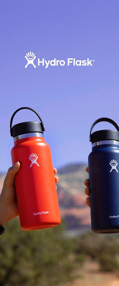 Dvi rankos laiko raudono ir tamsiai mėlyno Hydro Flask vandens butelius lauke, fonas – skaidrus mėlynas dangus ir tolumoje matoma akmenuota kraštovaizdis.