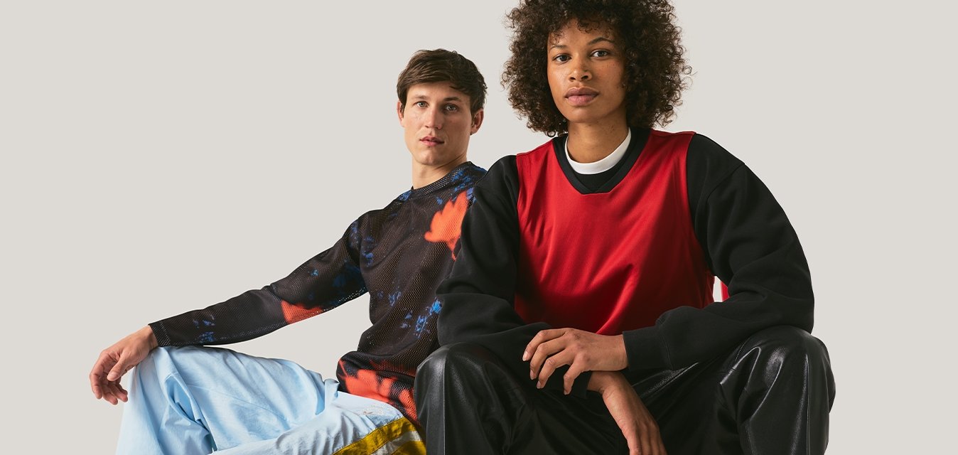 Dva mladí dospělí v běžném streetwearu, jeden sedí na červené stoličce a druhý sedí na podlaze opřený o jednoduché světlé pozadí.