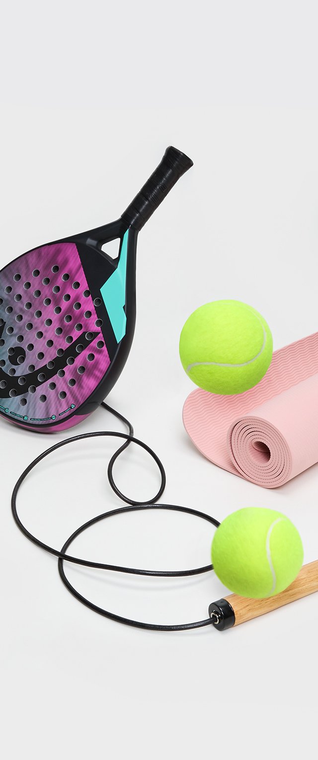 Racchetta da padel con una faccia texturizzata nera e viola, un accento teal neon, due palline da tennis verdi e un tappetino per yoga rosa arrotolato accanto a una corda per saltare.