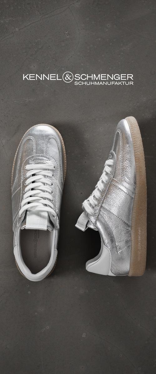 Zapatillas plateadas metálicas con parte superior de cuero texturizado, cordones blancos y suela de goma beige. Incluyen un detalle de logo en el lateral para la marca.